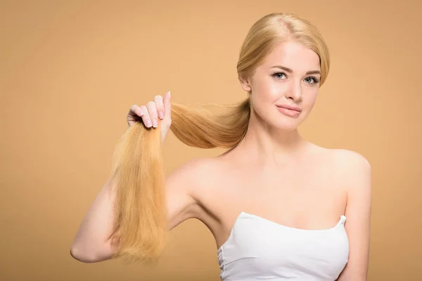 Hermosa joven rubia sosteniendo el pelo largo y sonriendo a la cámara aislada en beige - foto de stock