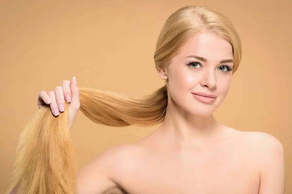 Attractive naked woman holding long blond hair and smiling at camera isolated on beige — Stock Photo