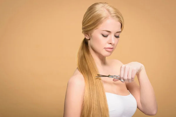 Junge blonde Frau hält Schere in der Hand und schneidet lange Haare isoliert auf beige — Stockfoto