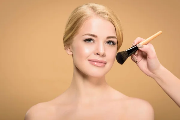 Recortado disparo de esteticista aplicación de maquillaje a la joven mujer desnuda mirando a la cámara aislada en beige - foto de stock