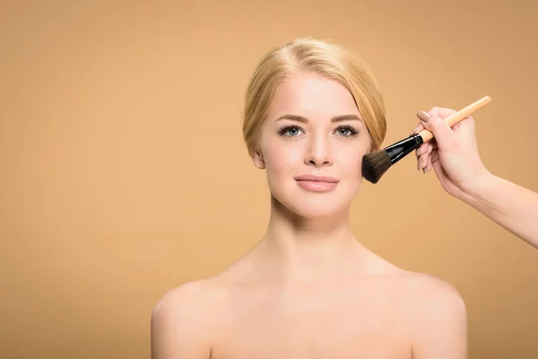 Recortado disparo de estilista aplicación de maquillaje con cepillo cosmético a la joven mujer desnuda mirando a la cámara aislada en beige - foto de stock