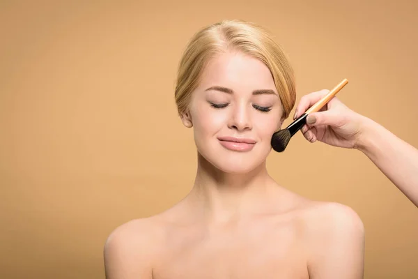 Plan recadré de styliste appliquant maquillage avec pinceau cosmétique à sourire femme nue isolée sur beige — Photo de stock