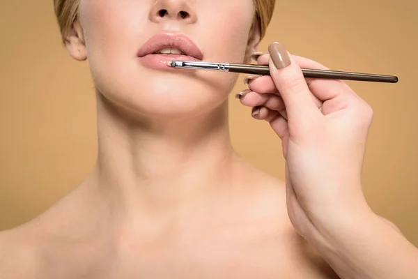 Abgeschnittene Aufnahme einer Stylistin mit Kosmetikpinsel, die Lippenstift auf eine schöne junge Frau aufträgt, isoliert auf Beige — Stockfoto