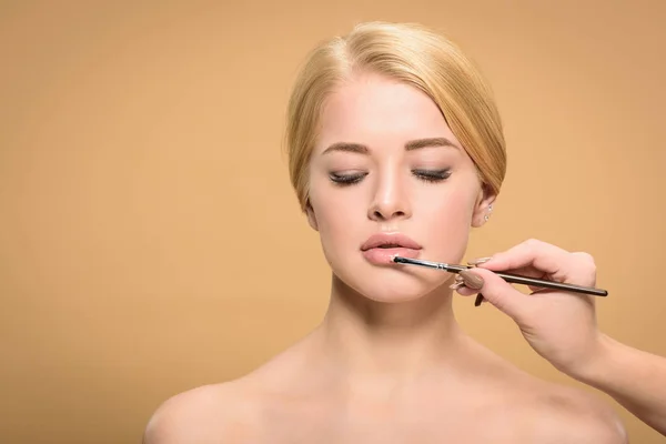 Abgeschnittene Aufnahme von Stylistin, die Kosmetikpinsel hält und Lippenstift auf schönes blondes Mädchen in Beige aufträgt — Stockfoto