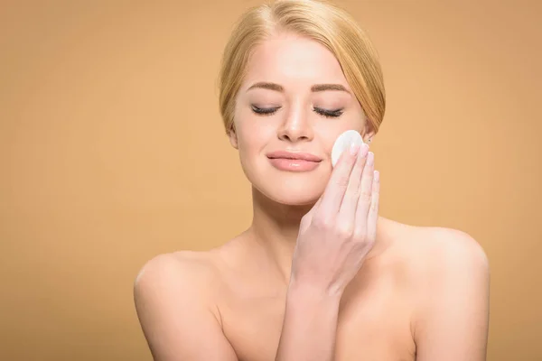 Bela mulher nua feliz com olhos fechados limpeza rosto com disco de algodão isolado no bege — Fotografia de Stock