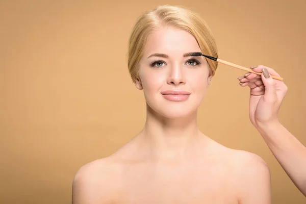 Stylish applying makeup on eyebrow to beautiful smiling naked woman isolated on beige — Stock Photo