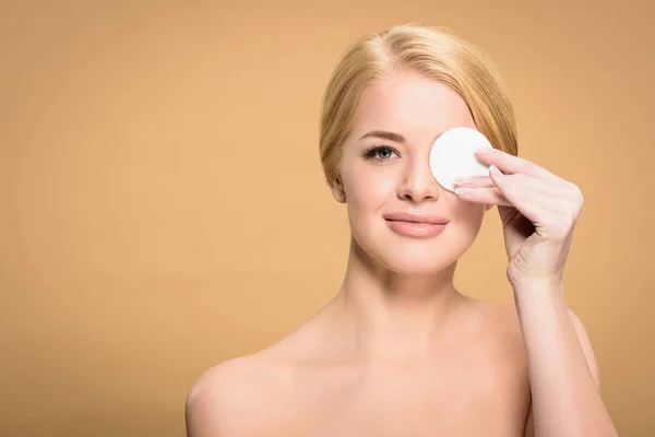 Attractive naked woman holding cotton disk near eye and smiling at camera isolated on beige — Stock Photo