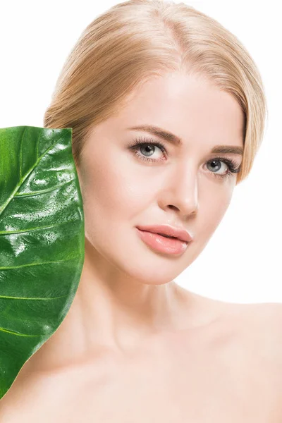 Привлекательная голая девушка с зеленым тропическим листом возле лица, смотрящая на камеру, изолированную на белом — стоковое фото