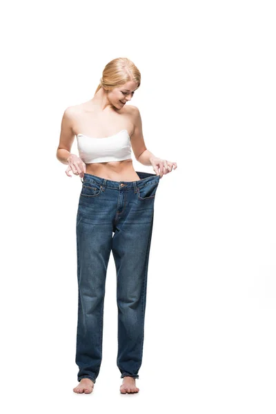 Piena vista lunghezza di sorridente ragazza magra che indossa jeans oversize e guardando verso il basso isolato su bianco — Foto stock