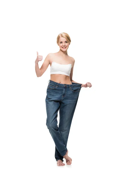 Mujer delgada feliz en pantalones vaqueros de gran tamaño mostrando el pulgar hacia arriba y sonriendo a la cámara aislada en blanco - foto de stock