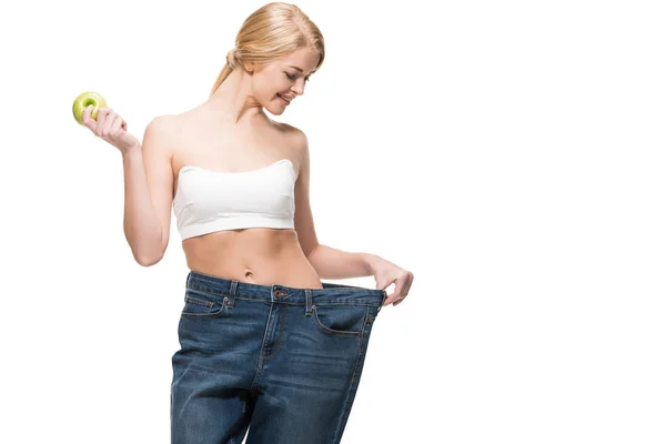 Schöne lächelnde junge Frau mit grünem Apfel und Blick auf übergroße Jeans isoliert auf weiß — Stockfoto