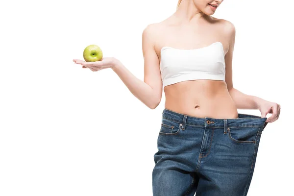 Обрезанный снимок улыбающейся худой женщины в огромных джинсах с зеленым яблоком, изолированным на белом — стоковое фото