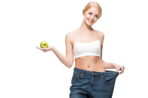 Beautiful slim girl in oversized jeans holding fresh apple and smiling at camera isolated on white — Stock Photo