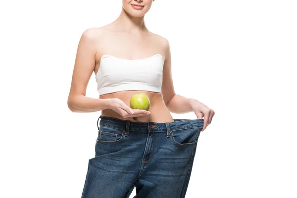 Corte tiro de sorrir jovem mulher em jeans oversized segurando maçã verde isolado no branco — Fotografia de Stock