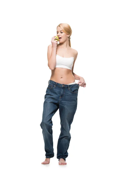 Beautiful young woman in oversized jeans eating green apple isolated on white — Stock Photo