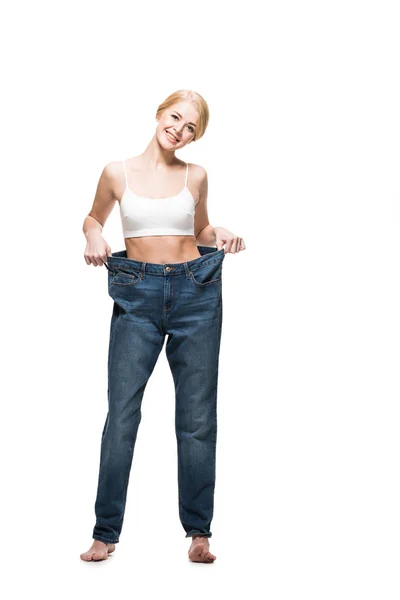 Full length view of happy young woman in oversized jeans smiling at camera isolated on white — Stock Photo