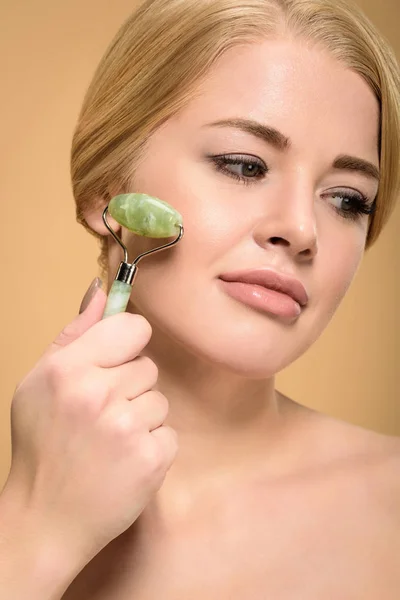 Atractiva joven mujer desnuda masajeando la cara con rodillo de jade y mirando hacia otro lado aislado en beige - foto de stock