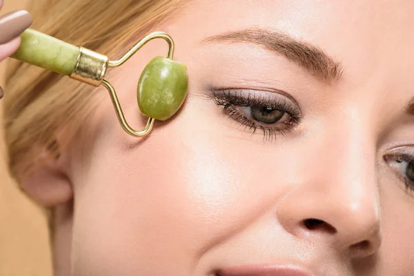 Vista ravvicinata di giovane donna massaggiante viso con rullo di giada isolato su beige — Foto stock