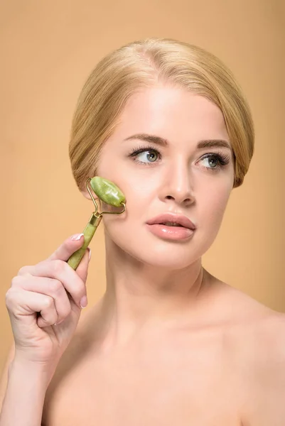 Attractive naked girl massaging face with jade roller and looking away isolated on beige — Stock Photo