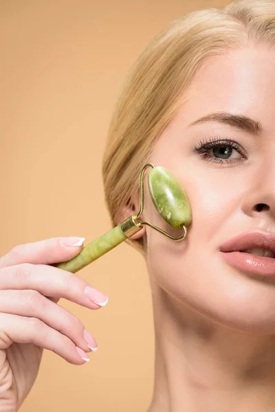 Plan recadré de jeune femme massant le visage avec un rouleau de jade et regardant la caméra isolée sur beige — Photo de stock