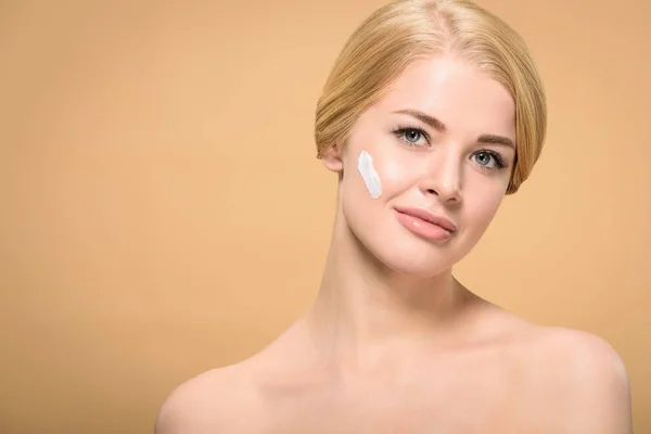 Attractive young woman with cosmetic cream on face smiling at camera isolated on beige — Stock Photo