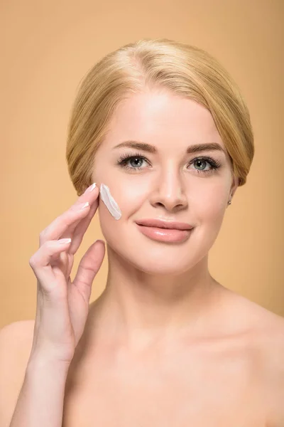 Attractive naked woman applying face cream and smiling at camera isolated on beige — Stock Photo