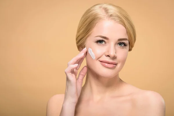 Beautiful naked woman applying face cream and smiling at camera isolated on beige — Stock Photo