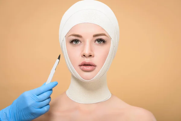 Cropped shot of doctor holding scalpel near woman with bandages over head isolated on beige — Stock Photo