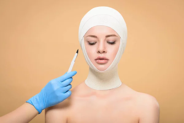 Coup de main en gant de latex tenant scalpel près de la femme avec des bandages sur la tête regardant vers le bas isolé sur beige — Photo de stock