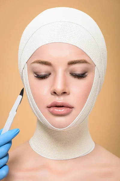 Cropped shot of hand in latex glove holding scalpel near woman with bandages over head isolated on beige — Stock Photo