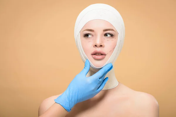 Doctor in latex glove touching face of young woman with bandages on head and correcting lines isolated on beige — Stock Photo