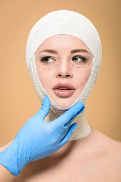 Plan recadré du médecin en gant de latex touchant le visage de la jeune femme avec des bandages sur la tête et des lignes correctrices isolées sur beige — Photo de stock