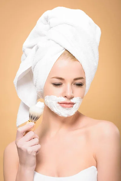Mulher com toalha na cabeça e creme de barbear no rosto segurando escova de barbear e olhando para baixo isolado no bege — Fotografia de Stock