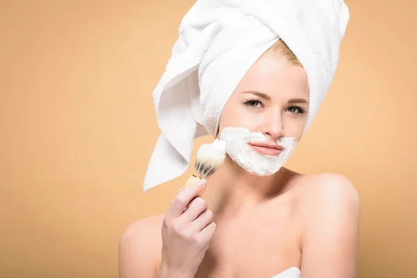 Jeune femme avec serviette sur la tête appliquer de la mousse avec brosse à raser isolé sur beige — Photo de stock