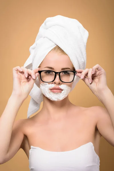 Donna con asciugamano sulla testa e crema da barba sul viso che regola gli occhiali e guardando la fotocamera isolata sul beige — Foto stock