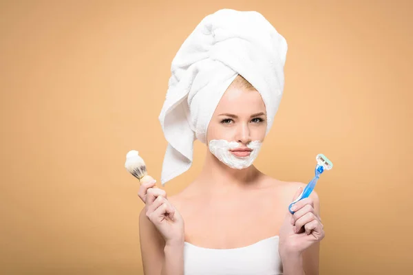 Young woman with towel on head and shaving cream on face holding razor and shaving brush isolated on beige — Stock Photo
