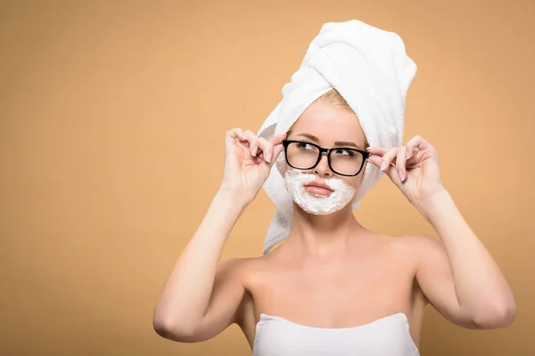 Молода жінка з рушником на голові і кремом для гоління на обличчі, що регулює окуляри і дивиться ізольовано на бежевий — стокове фото
