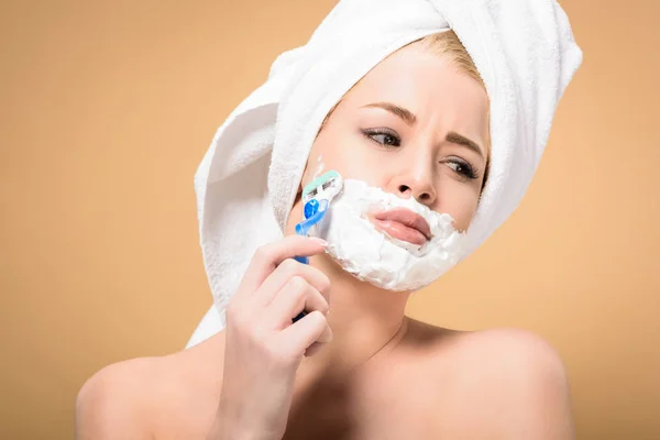 Mujer joven desnuda con toalla en la cabeza afeitándose la cara con afeitadora y mirando hacia otro lado aislado en beige - foto de stock