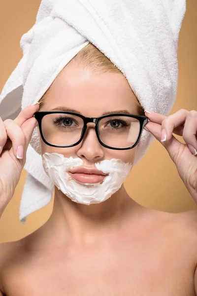 Beautiful naked with with towel on head and shaving cream on face adjusting eyeglasses isolated on beige — Stock Photo