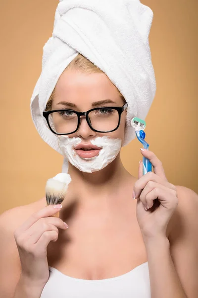 Mujer joven con toalla en la cabeza sosteniendo afeitadora y cepillo de afeitar aislado en beige - foto de stock