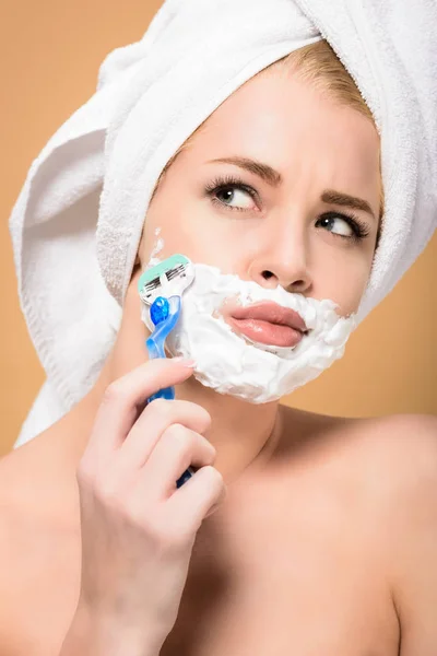 Young naked woman with towel on head shaving face with razor isolated on beige — Stock Photo