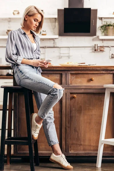 Atractiva mujer usando teléfono inteligente y sentado en la silla - foto de stock