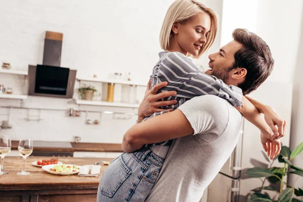 Novio guapo y novia atractiva abrazando y sonriendo en la cocina - foto de stock