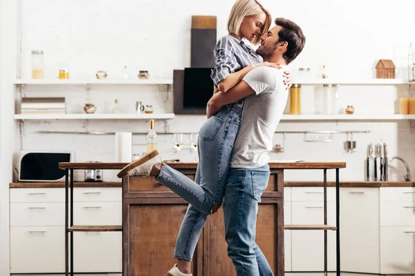 Novio guapo y novia atractiva abrazo y de pie en la cocina - foto de stock