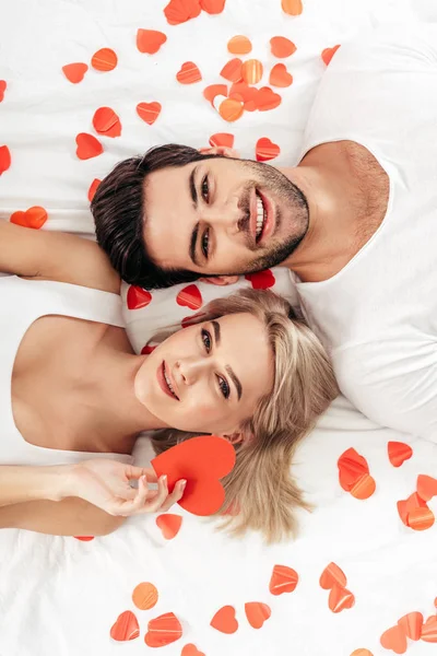 Top view of attractive girlfriend and handsome boyfriend smiling at camera and laying near heart signs on Valentine 's day — стоковое фото