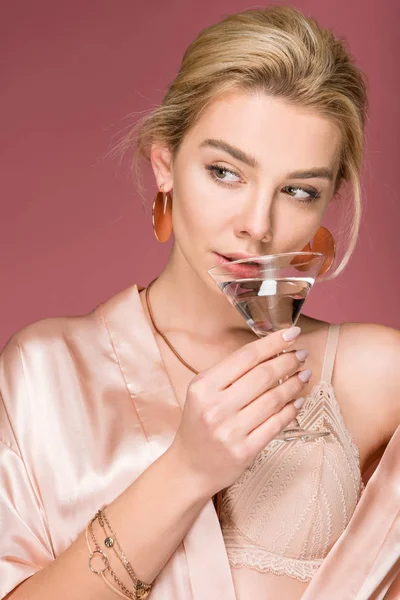Привлекательная молодая женщина в шелковом халате с коктейльным бокалом, изолированная на розовом — стоковое фото