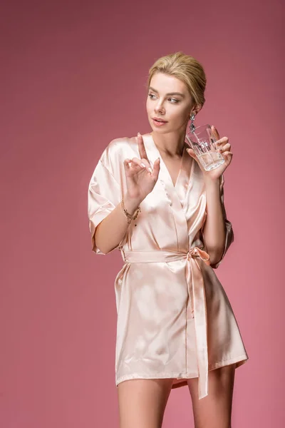 Chica atractiva en túnica de seda gesto y celebración de vidrio con cóctel, aislado en rosa - foto de stock