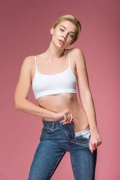Красива блондинка позує в джинсах і білому бюстгальтері, ізольована на рожевому — стокове фото