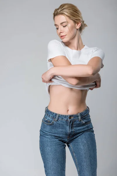 Молода жінка в джинсах і білій футболці роздягається ізольовано на сірому — стокове фото