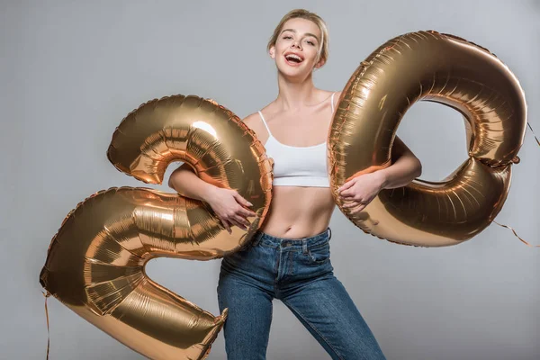 Excited beautiful girl in jeans and white bra posing with 20 golden balloons, isolated on grey — Stock Photo
